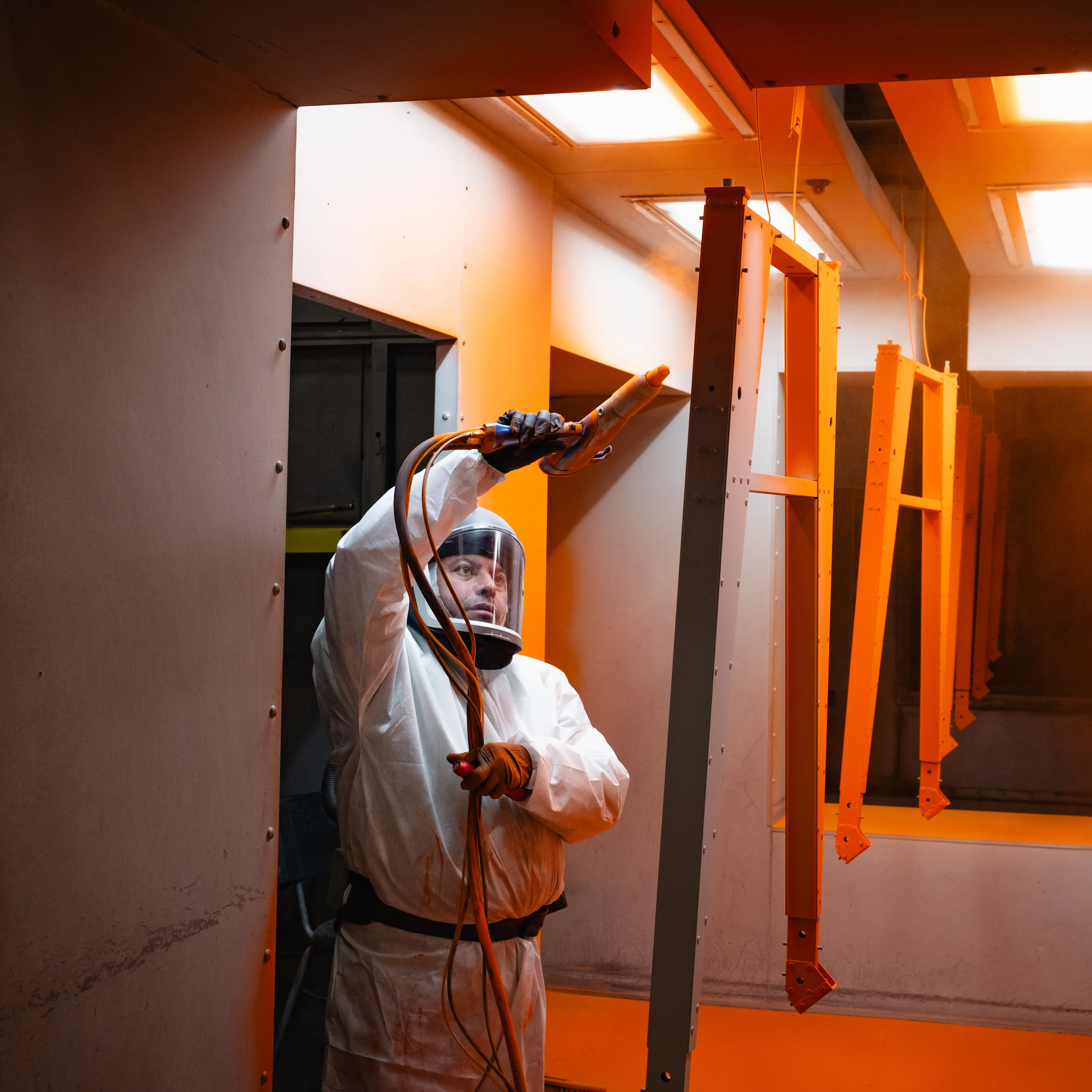 In an industrial setting with orange lighting, a person in protective gear showcases exceptional craftsmanship as they spray-paint metal frames, perfecting the art of custom modular cabinetry.